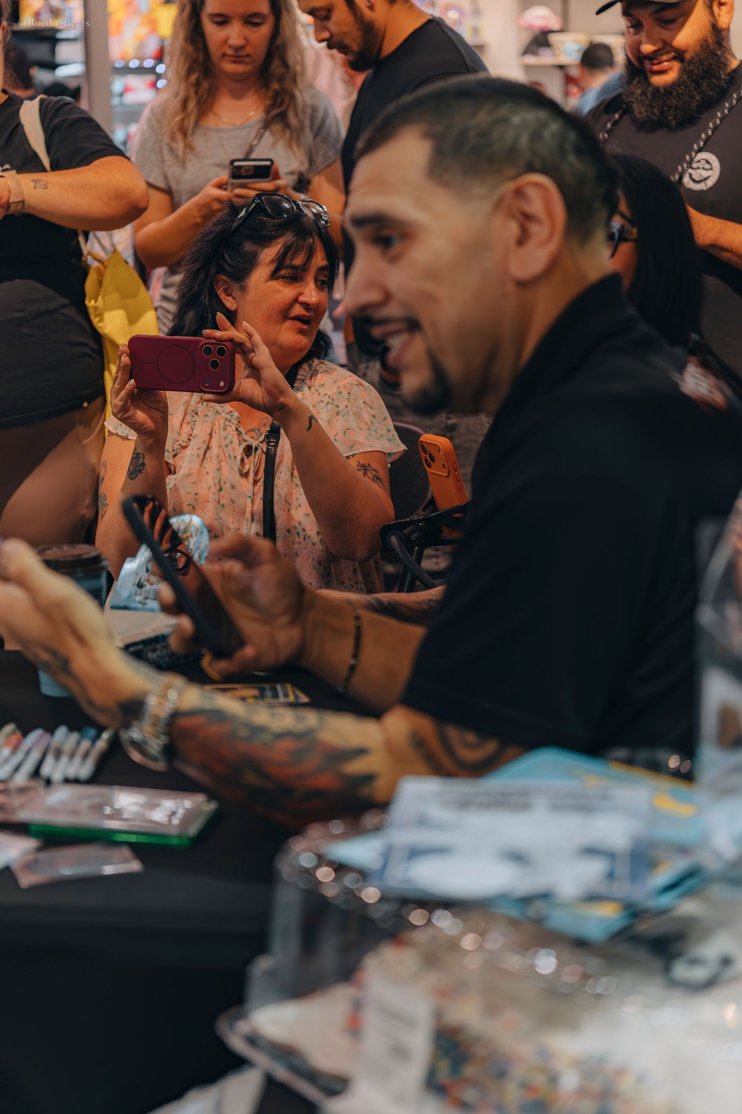 Tattooed man smiles as fan photographs him at trading card event.