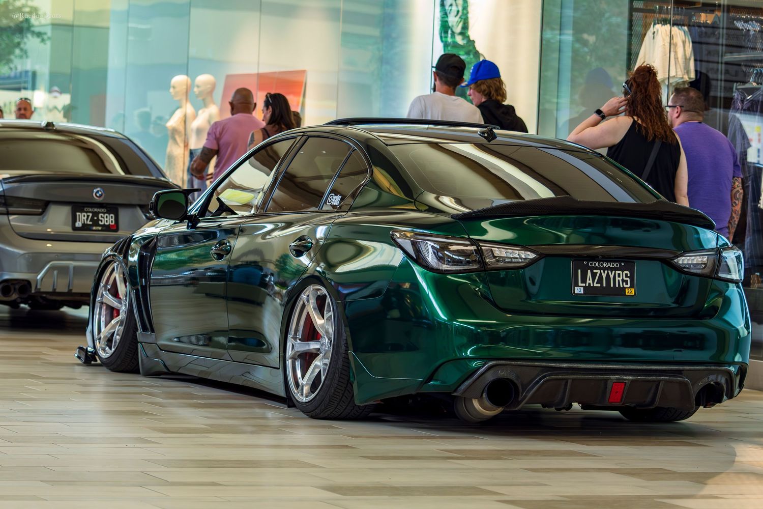 Lowered dark green Infiniti Q50 with red wheels and LAZYRS license plate at indoor car show.