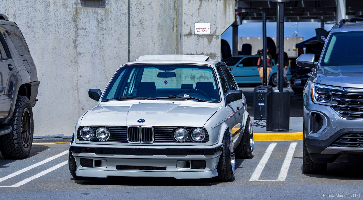 A silver BMW E30 with a front lip spoiler driving through a parking area at a car show.