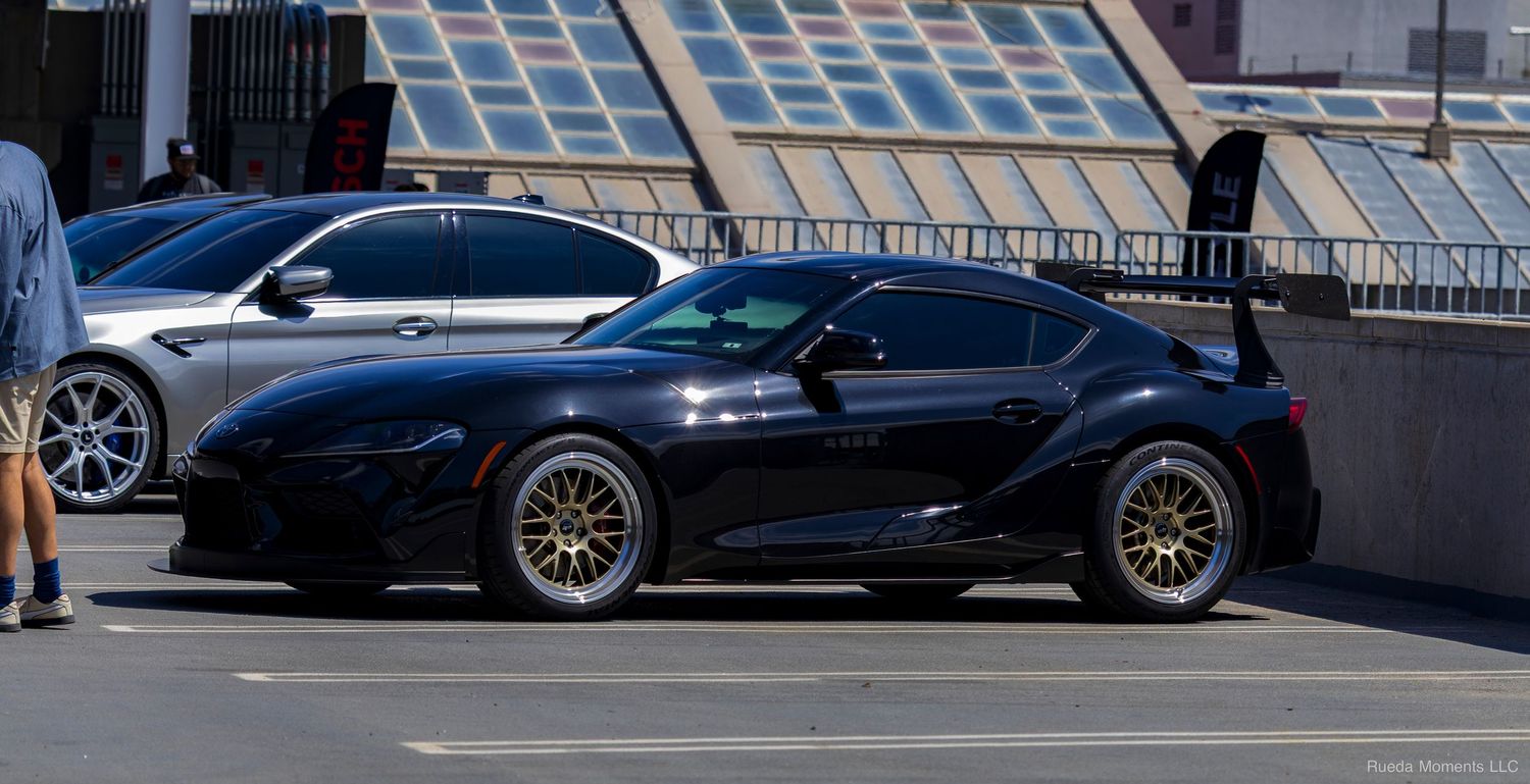 Black Toyota GR Supra with wide body kit and gold wheels parked on urban street.