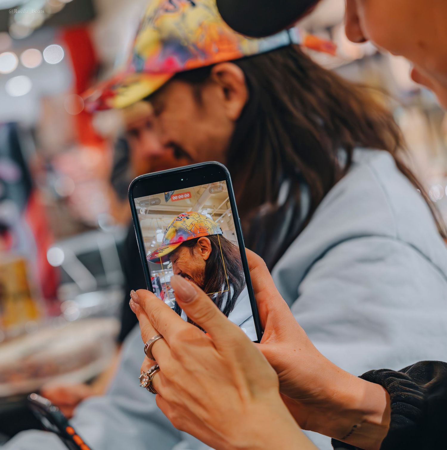 Person photographing a man in colorful cap with smartphone at a trading card signing event.
