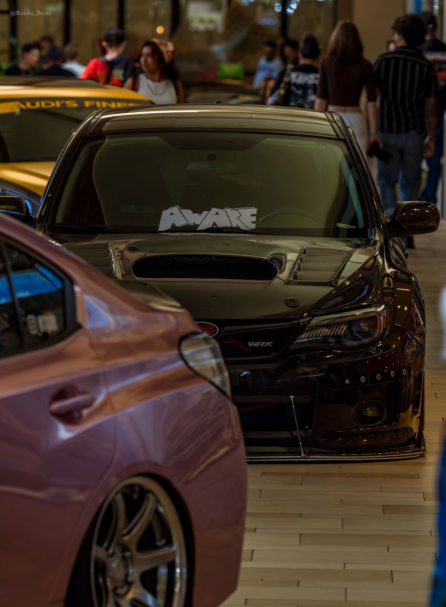 Dark Subaru WRX STI with carbon hood and AWARE windshield banner displayed at indoor mall car show.