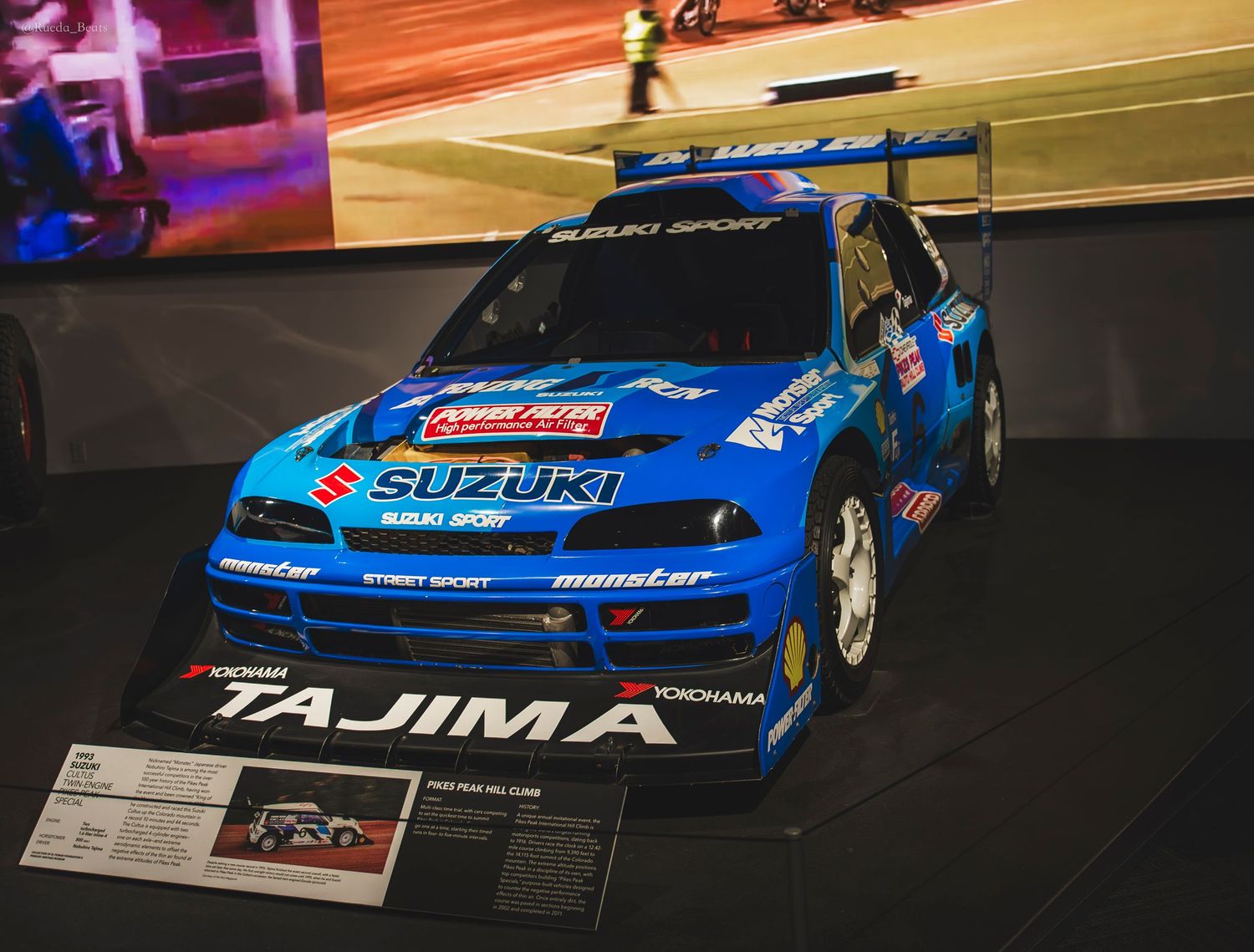 Blue Suzuki Tajima rally car number 16 on display at an indoor motorsport exhibition.