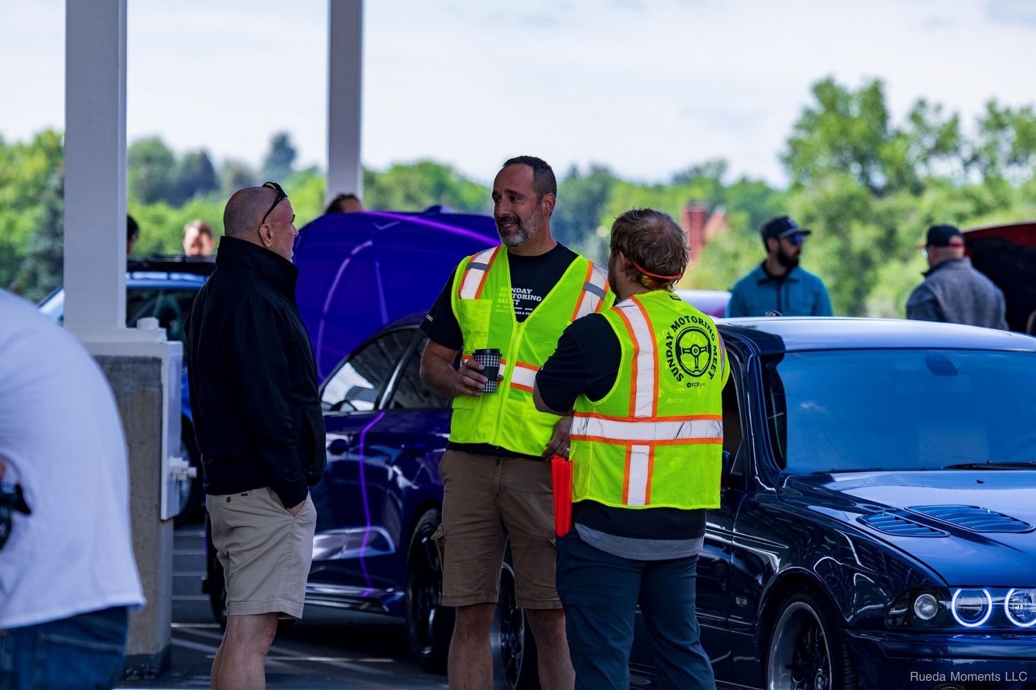 Two event marshals in yellow safety vests reviewing documents at an outdoor car show.