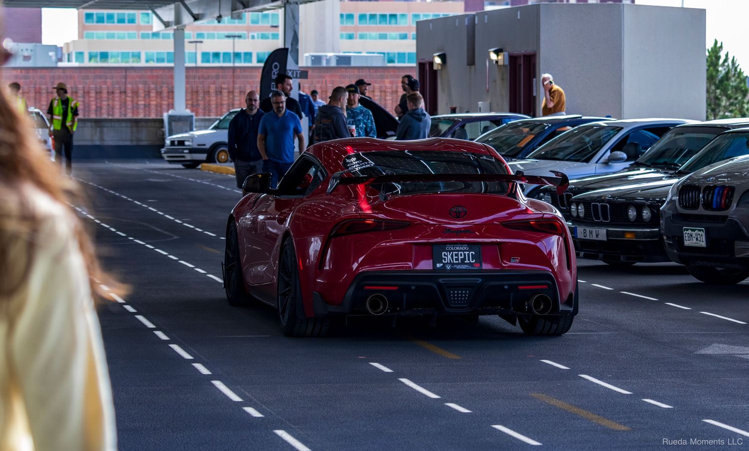 Red Toyota GR Supra driving through an outdoor automotive car show event at dusk.