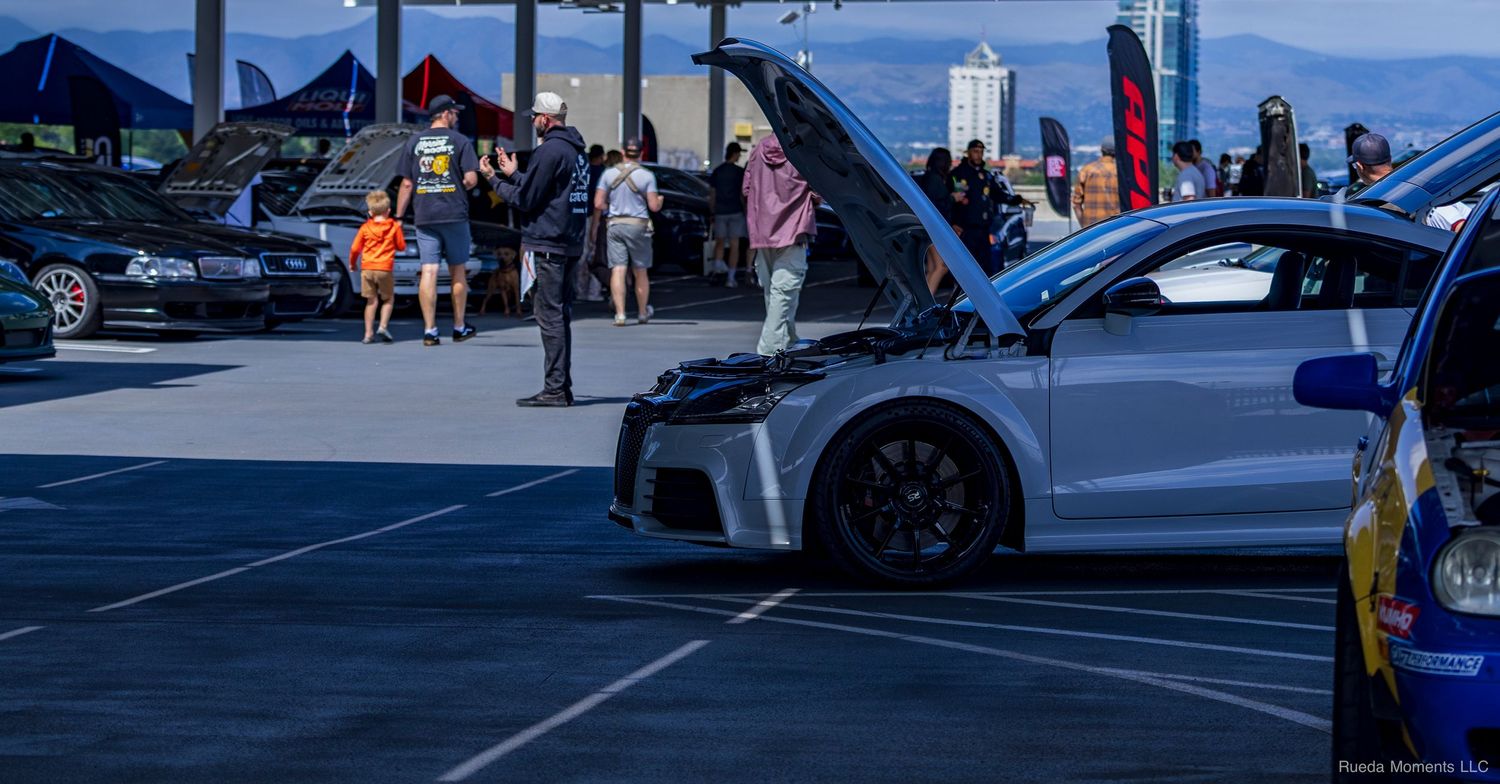 A silver sports car with hood open displayed at an outdoor automotive event near a city.