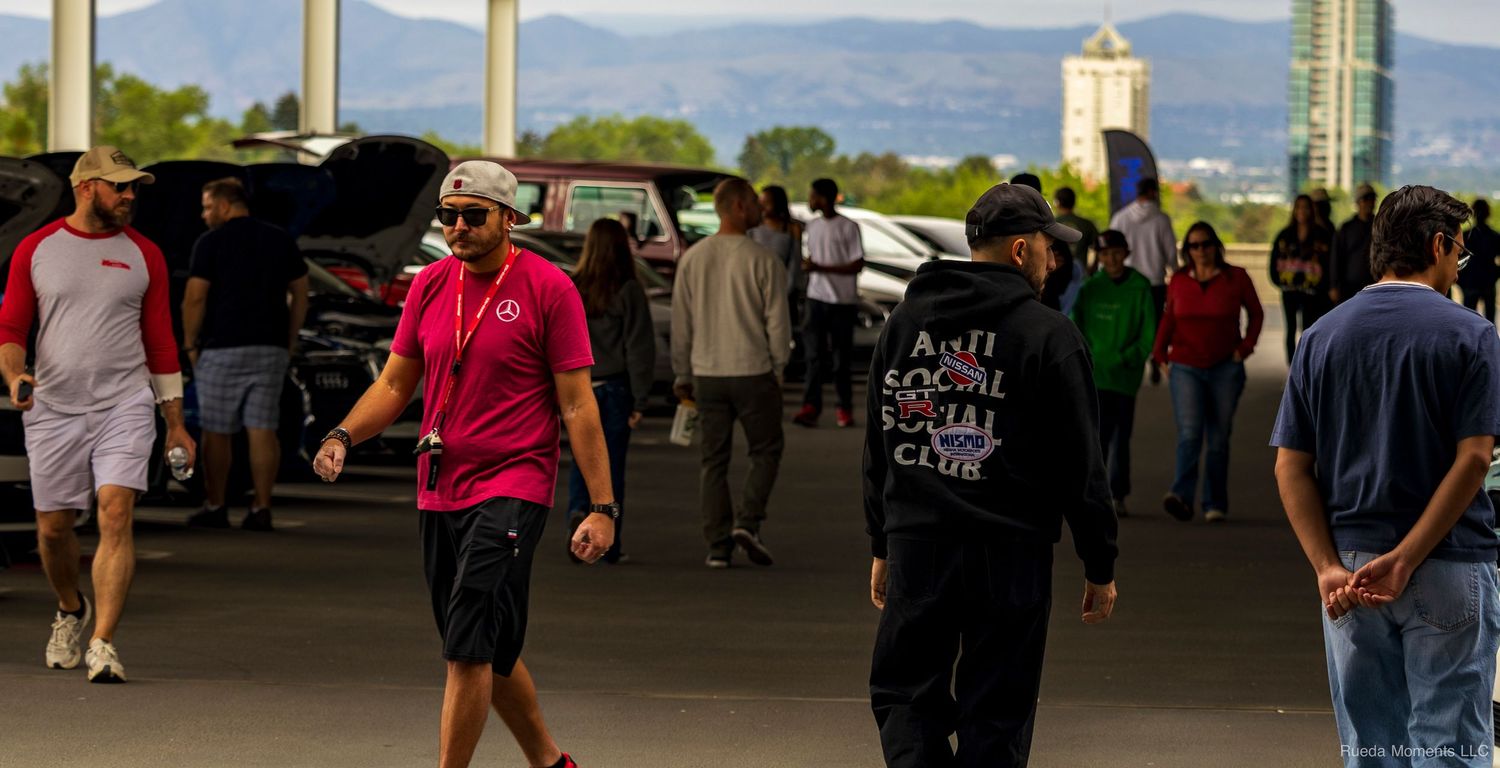 Car enthusiasts walking through a sunny outdoor automotive meet with mountains in background.