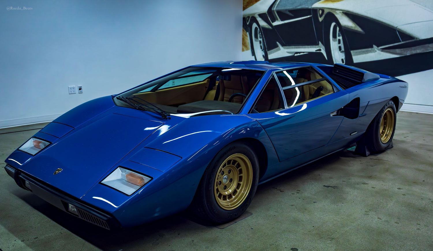 Blue Lamborghini Countach classic supercar with gold wheels displayed in a modern automotive museum.