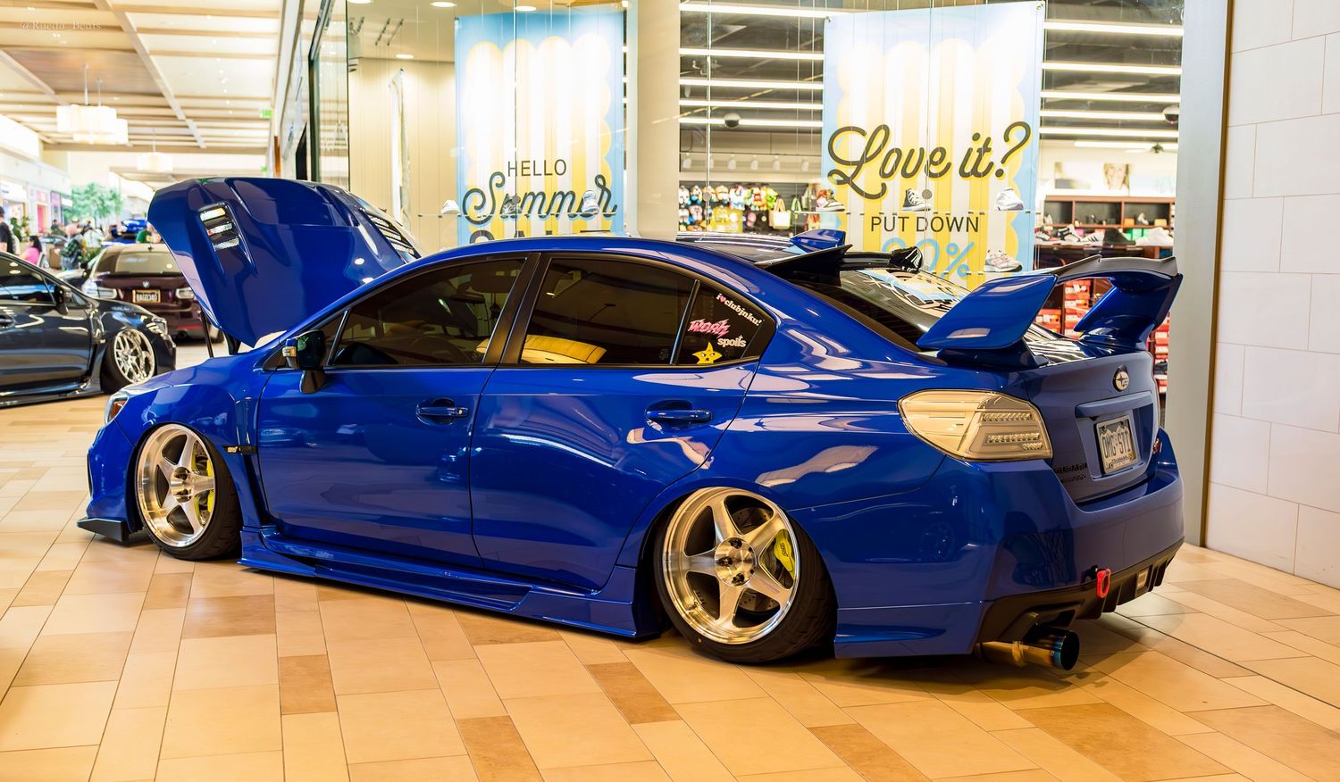 Blue Subaru WRX STI widebody on gold wheels with open hood at mall car show.