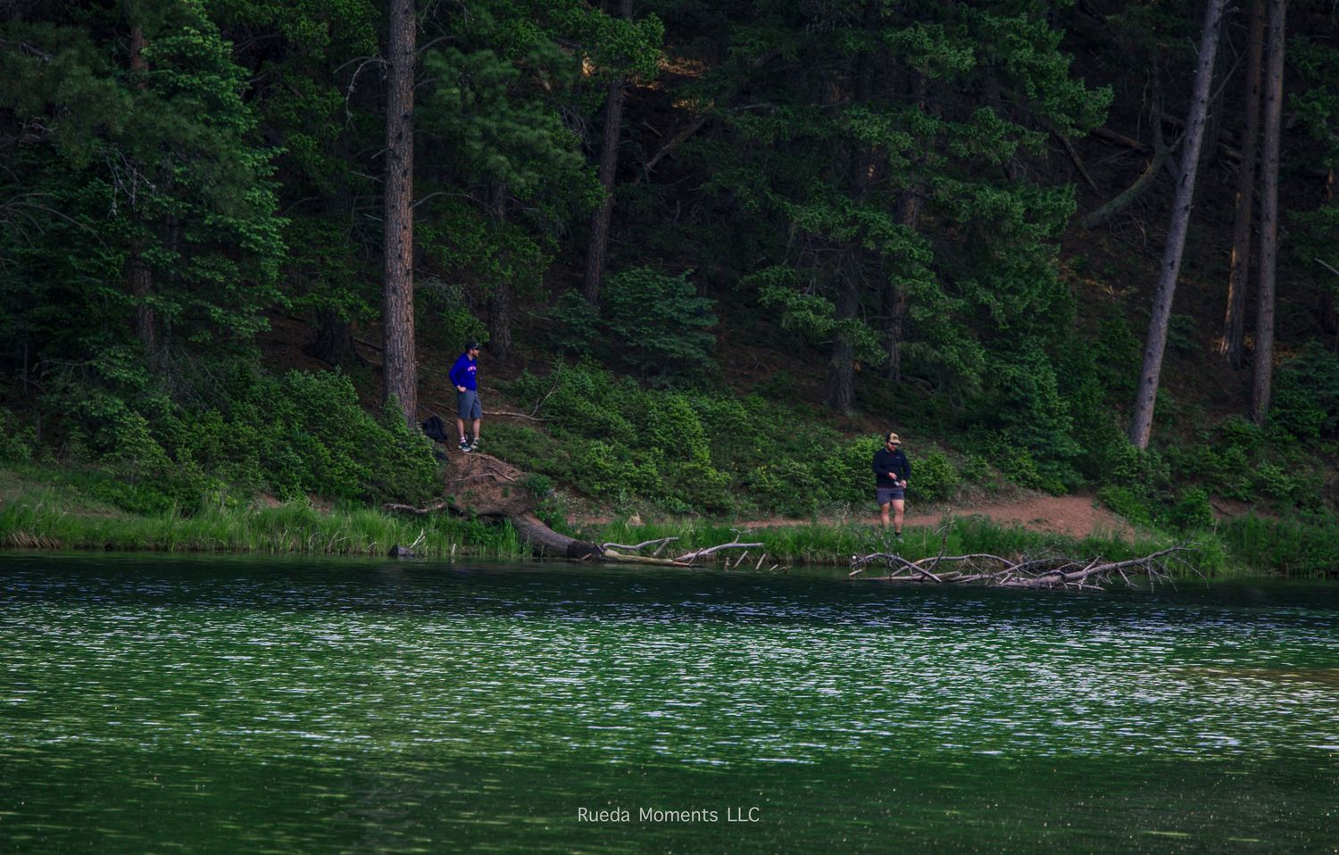 Two people fishing along the forested shoreline of a calm green mountain lake at dusk.