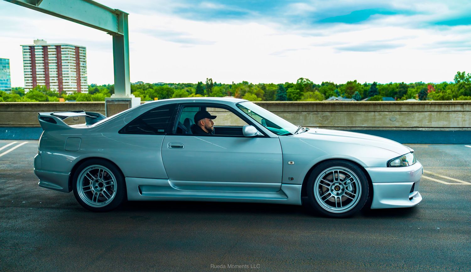 Silver Nissan Skyline R33 GT-R with BBS wheels driving on an elevated highway overpass.