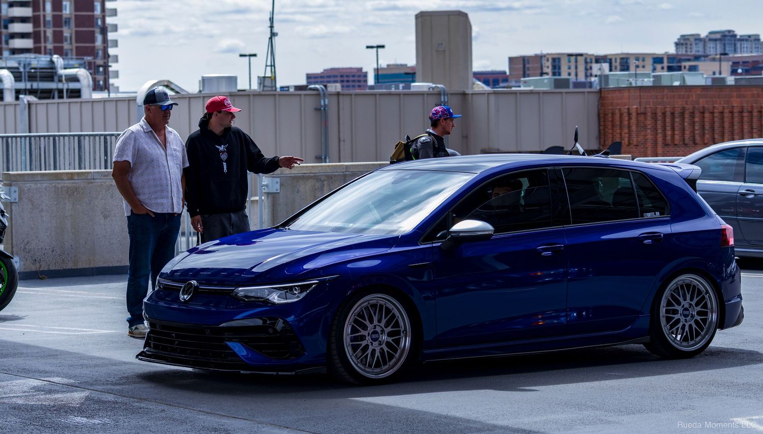 A Lapiz Blue Volkswagen Golf R Mk8 with three enthusiasts gathered around it at a car meet.