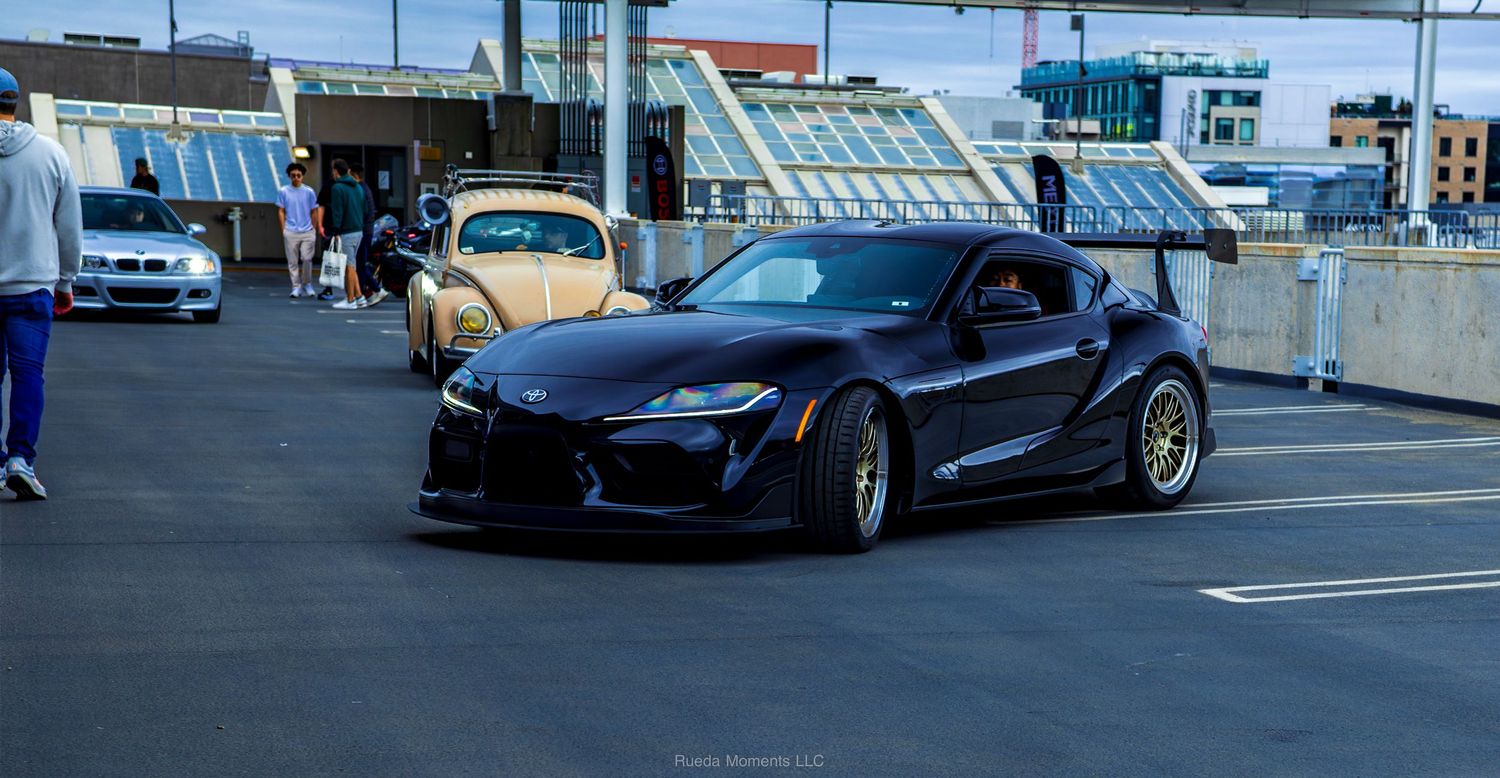 Modified black Toyota GR Supra with wide body kit at a car meet.
