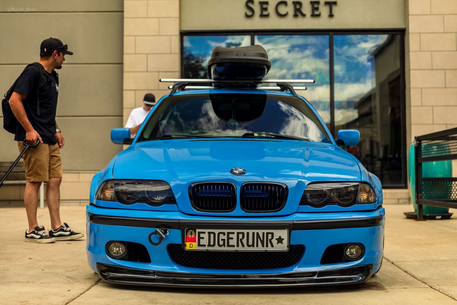 Laguna Seca Blue BMW E46 with roof box and EDGERUNR plate parked in front of a retail store.