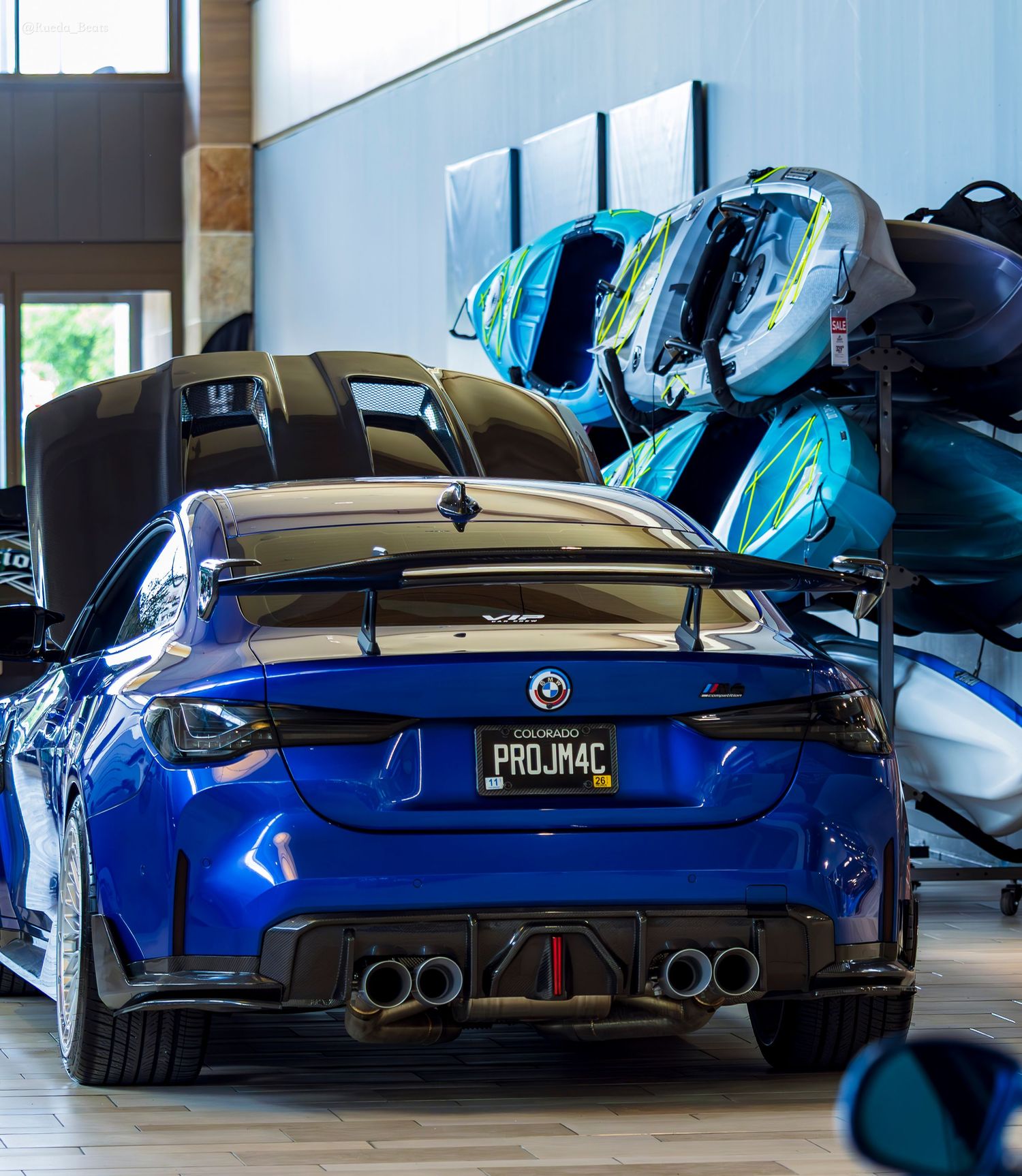 Blue BMW M4 with PROJM4C vanity plate and large rear spoiler displayed at indoor car show.