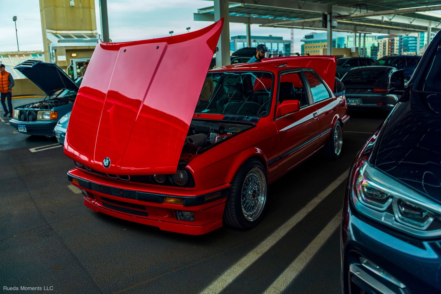 Classic red BMW E30 with hood open displaying engine bay at an outdoor car show.