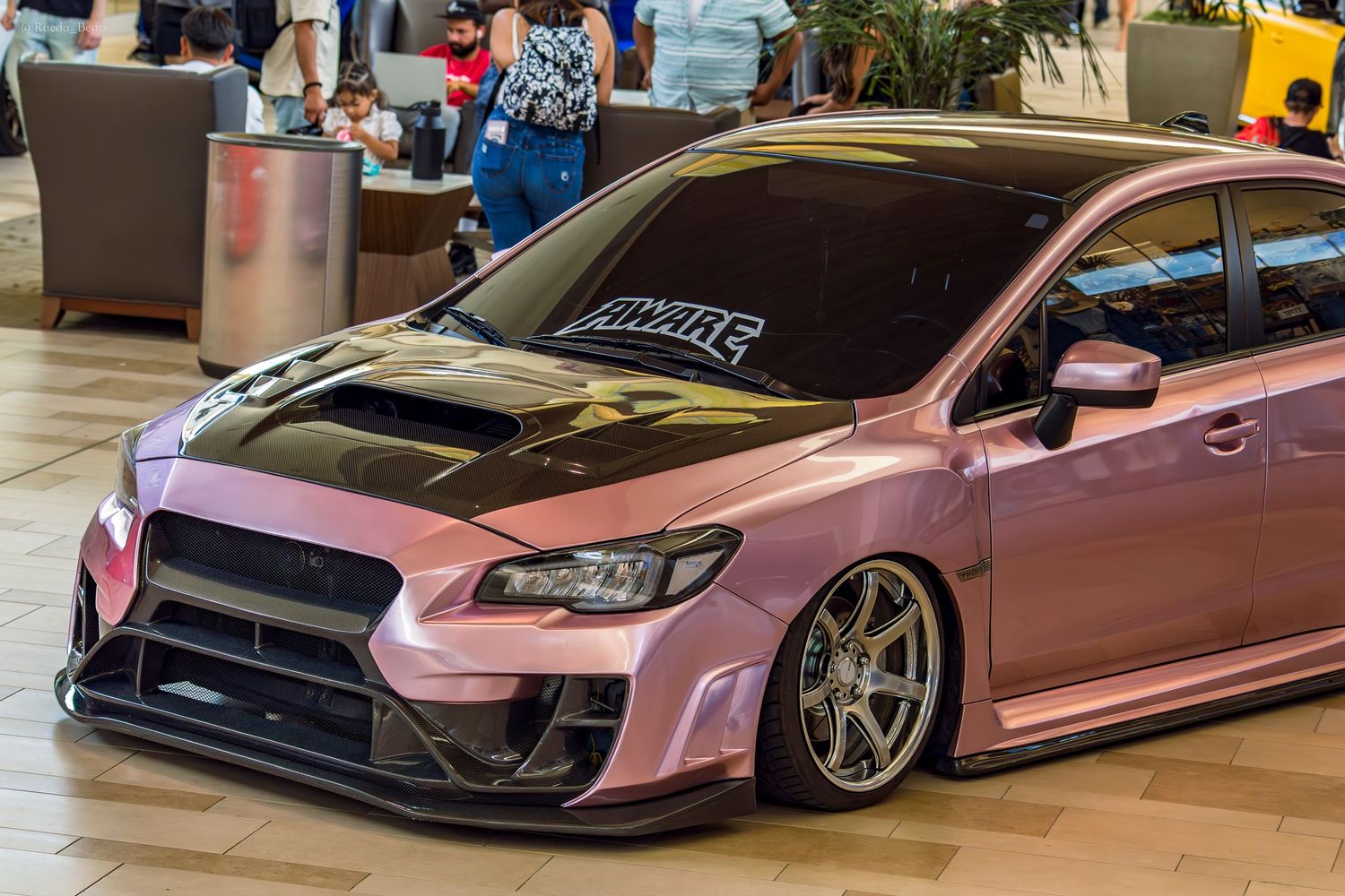 Widebody pink Subaru WRX STI with carbon hood and AWARE windshield banner at indoor car show.