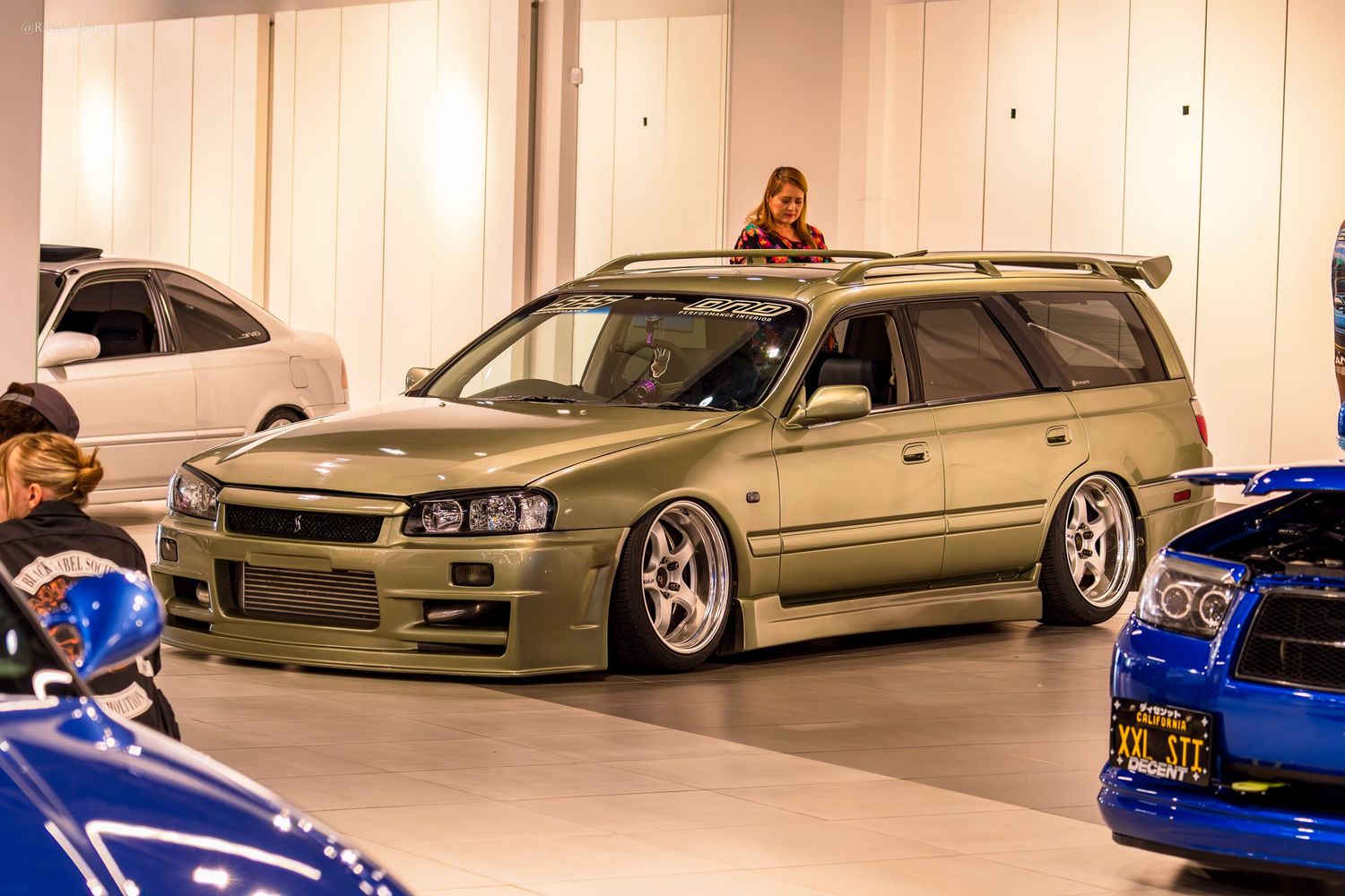 Stanced gold Nissan Stagea wagon on deep dish wheels at an indoor car show display.