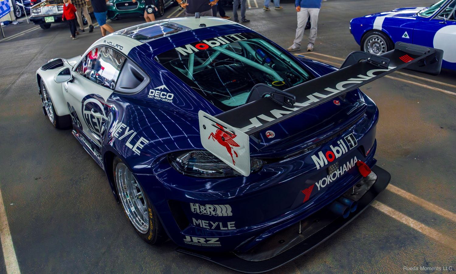 Blue racing Porsche 911 GT3 with sponsor decals displayed at an indoor car show.