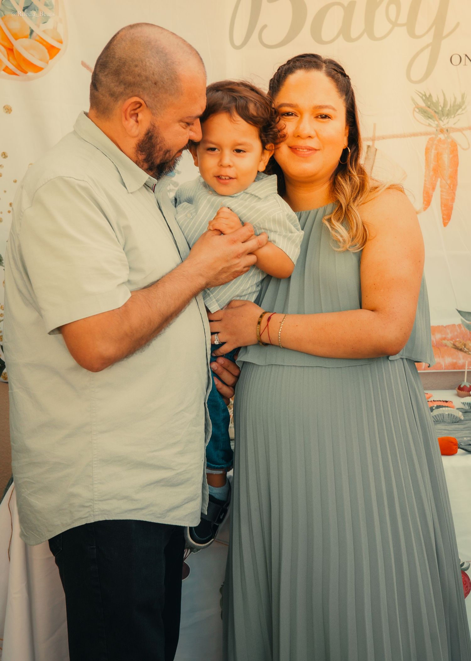 Happy family at baby shower with father holding toddler beside pregnant mother in sage green dress.