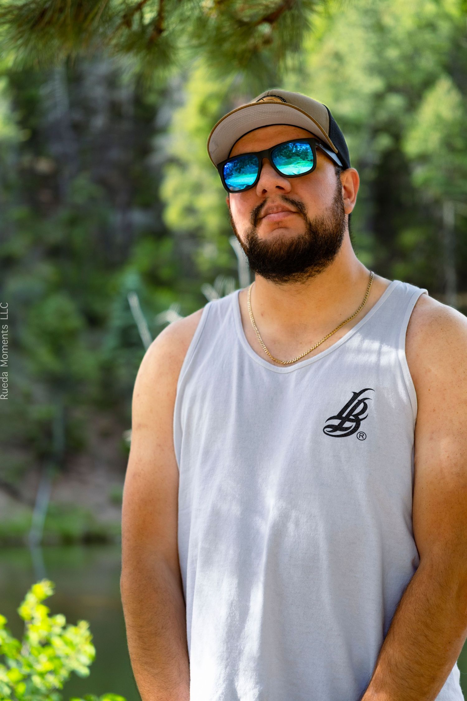 Bearded man wearing blue mirrored sunglasses and LB tank top poses outdoors near green trees.