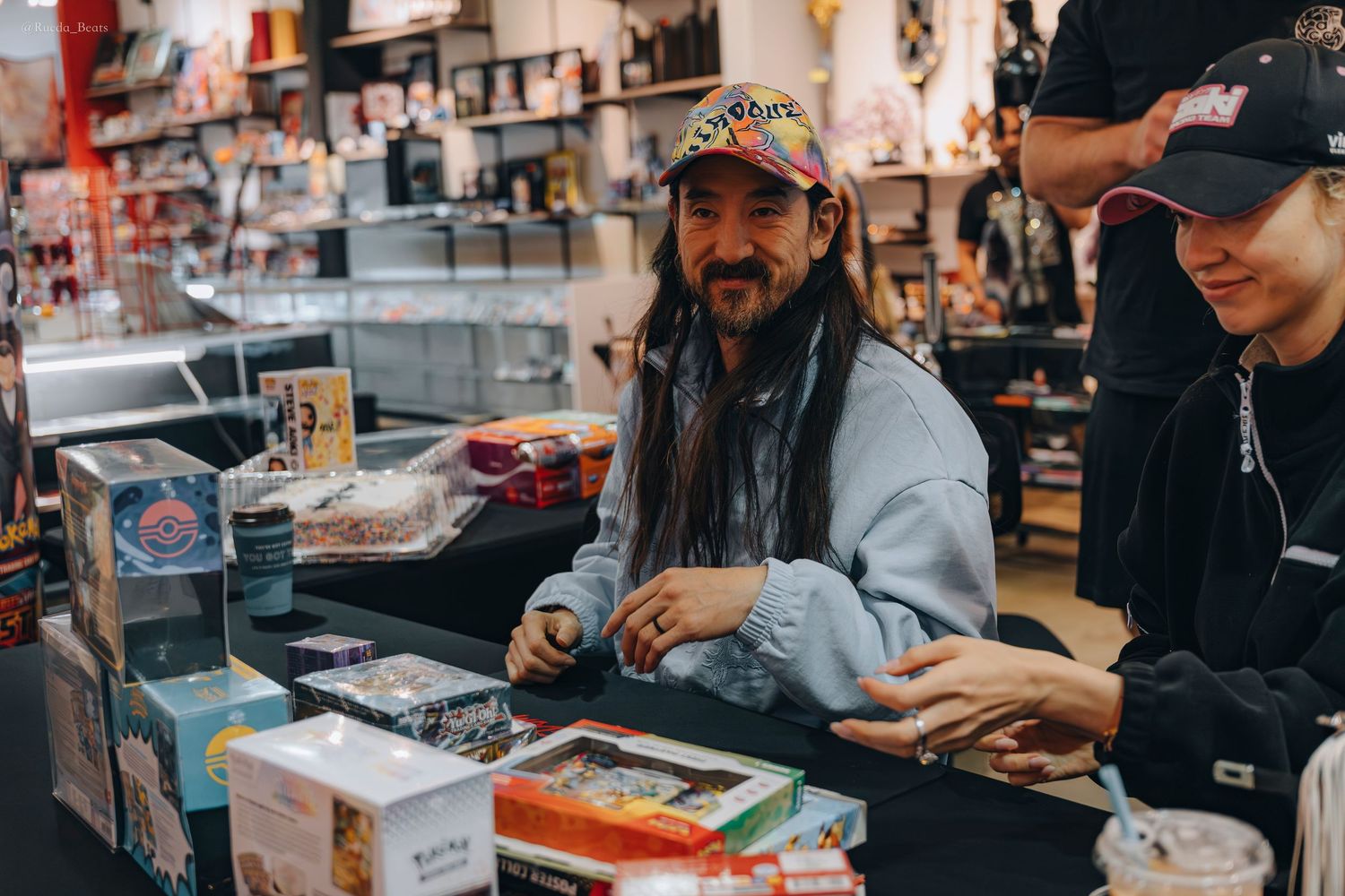 Smiling man in colorful cap sitting at Nerdz Kingdom store counter with trading cards.
