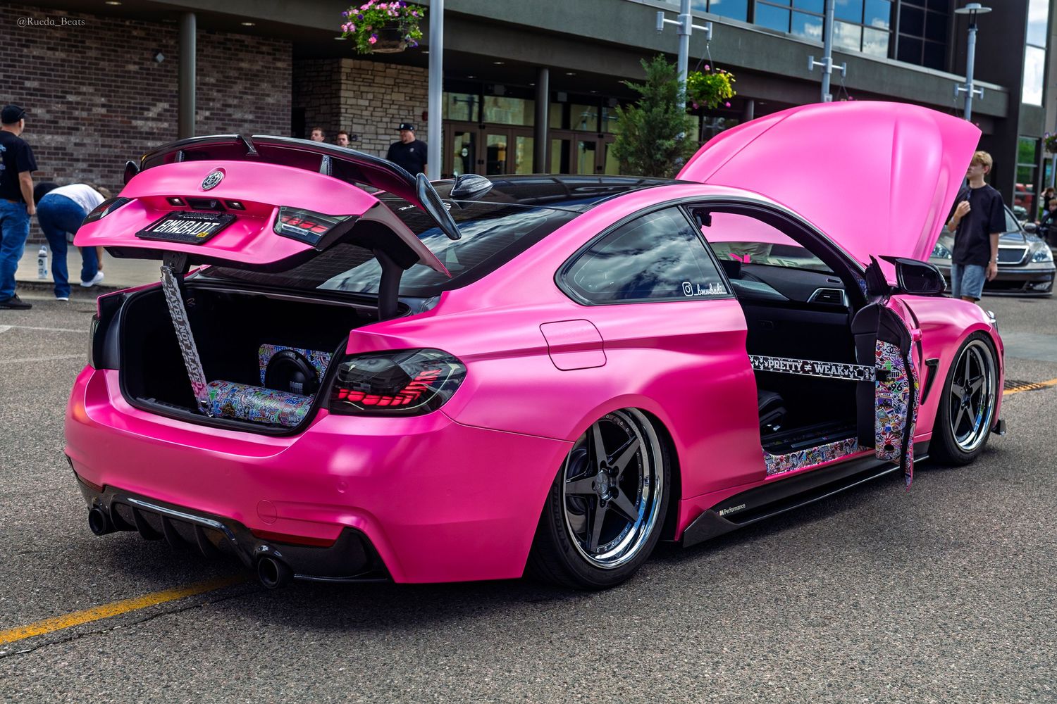 Hot pink widebody BMW M4 with open doors and trunk displayed at a car show.