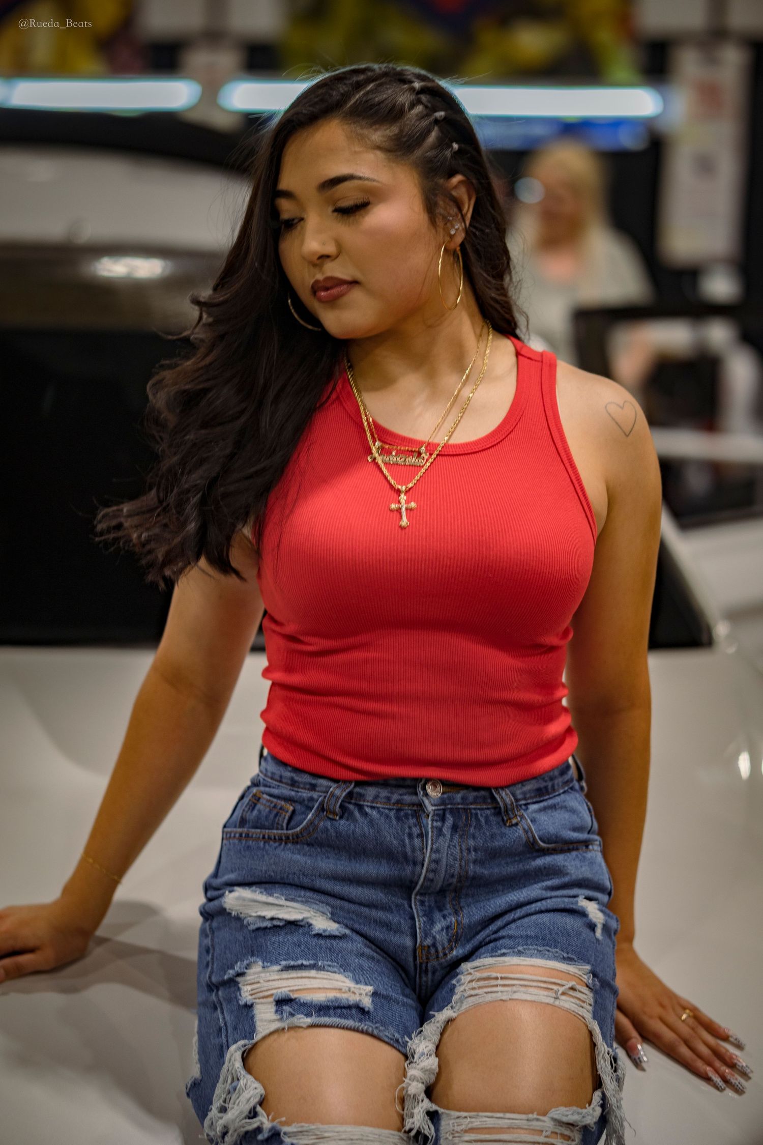 Young woman in red tank top and ripped denim shorts wearing gold cross necklace posing near a white car.