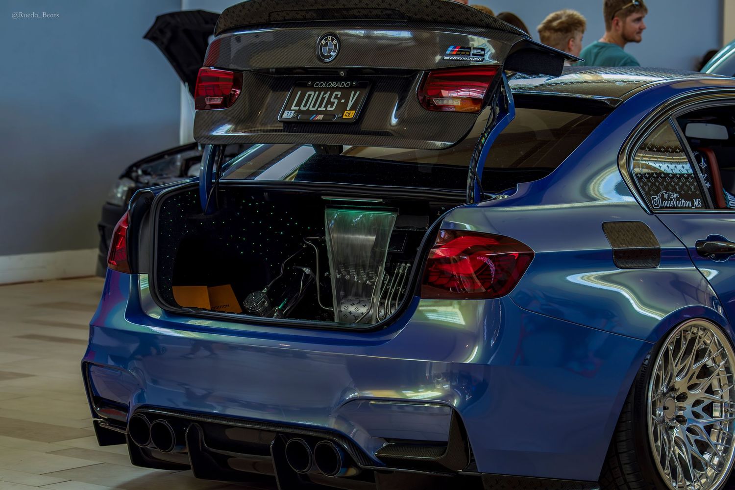 Blue BMW M3 with custom trunk audio setup and LOUIS V vanity plate at indoor car show.
