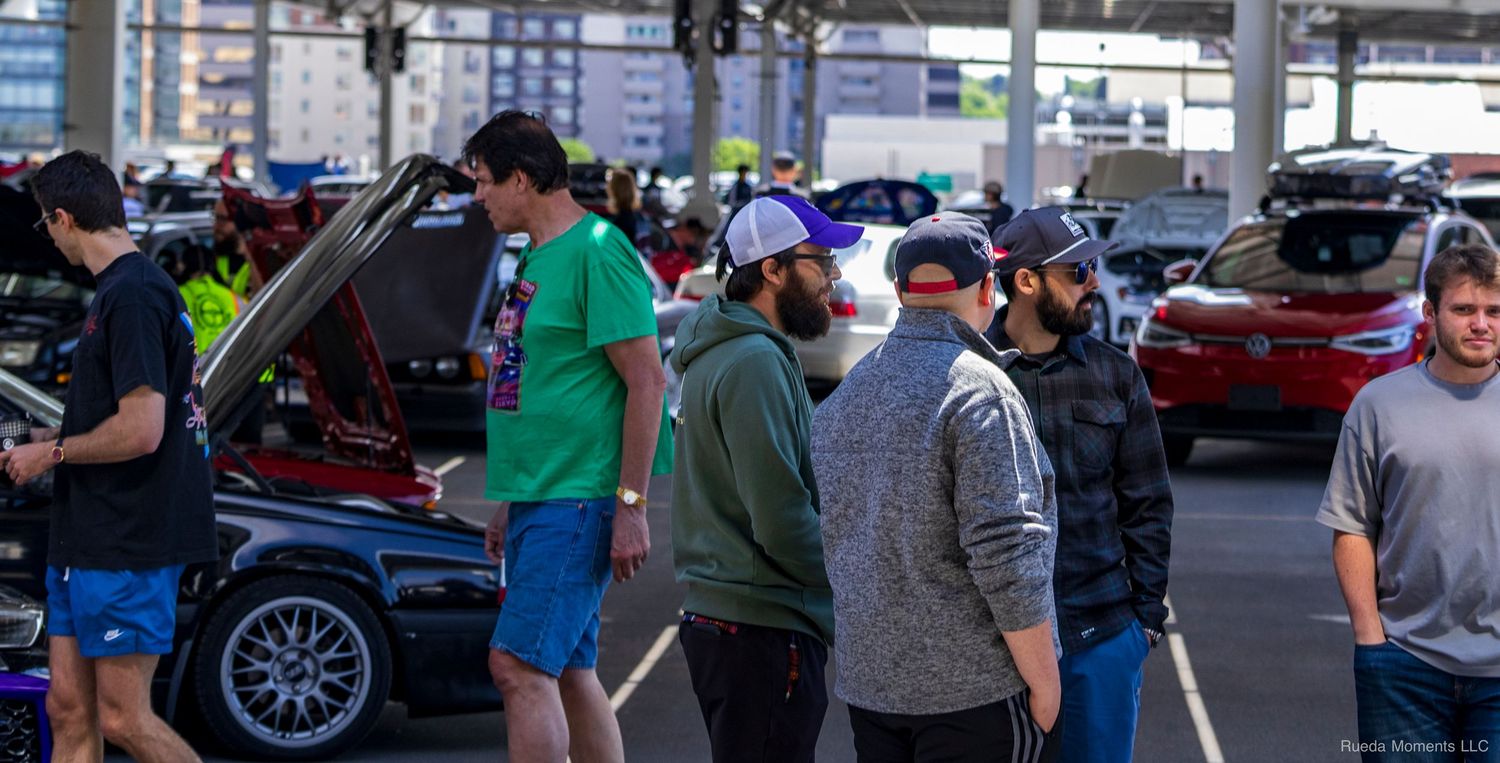 Car enthusiasts gather at an outdoor auto show, inspecting vehicles with hoods open in a busy parking lot.