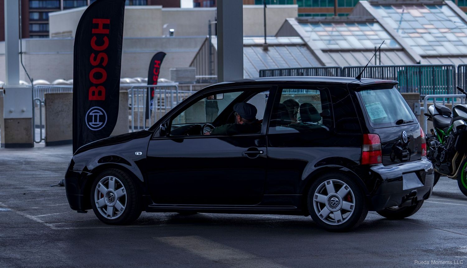 A black Volkswagen Lupo hatchback parked near a Bosch banner at a winter car meet.