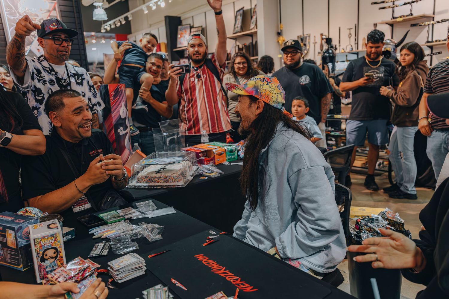 Excited crowd cheering at a man seated at a trading card signing table.