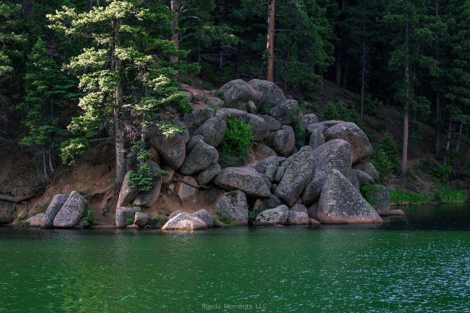 Large granite boulders cluster along the emerald green shoreline surrounded by tall pine trees in a mountain lake.