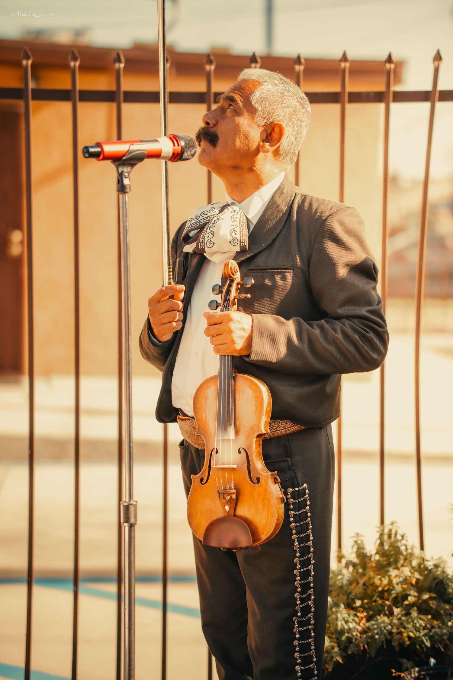 Mariachi musician in traditional black suit holding violin while singing passionately into microphone outdoors.