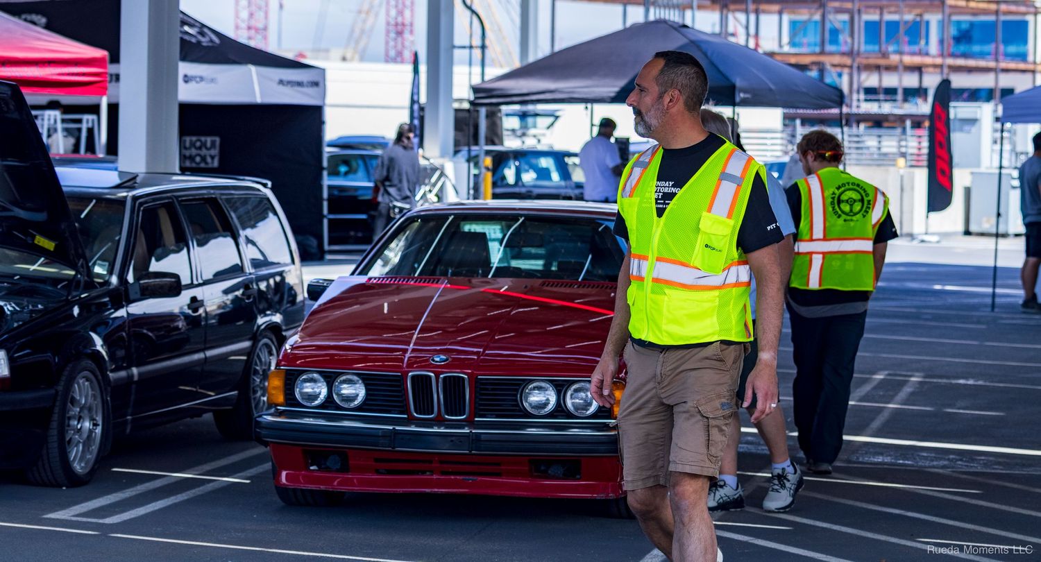 Man in yellow safety vest walking past a lowered red vintage BMW at an outdoor car show event.