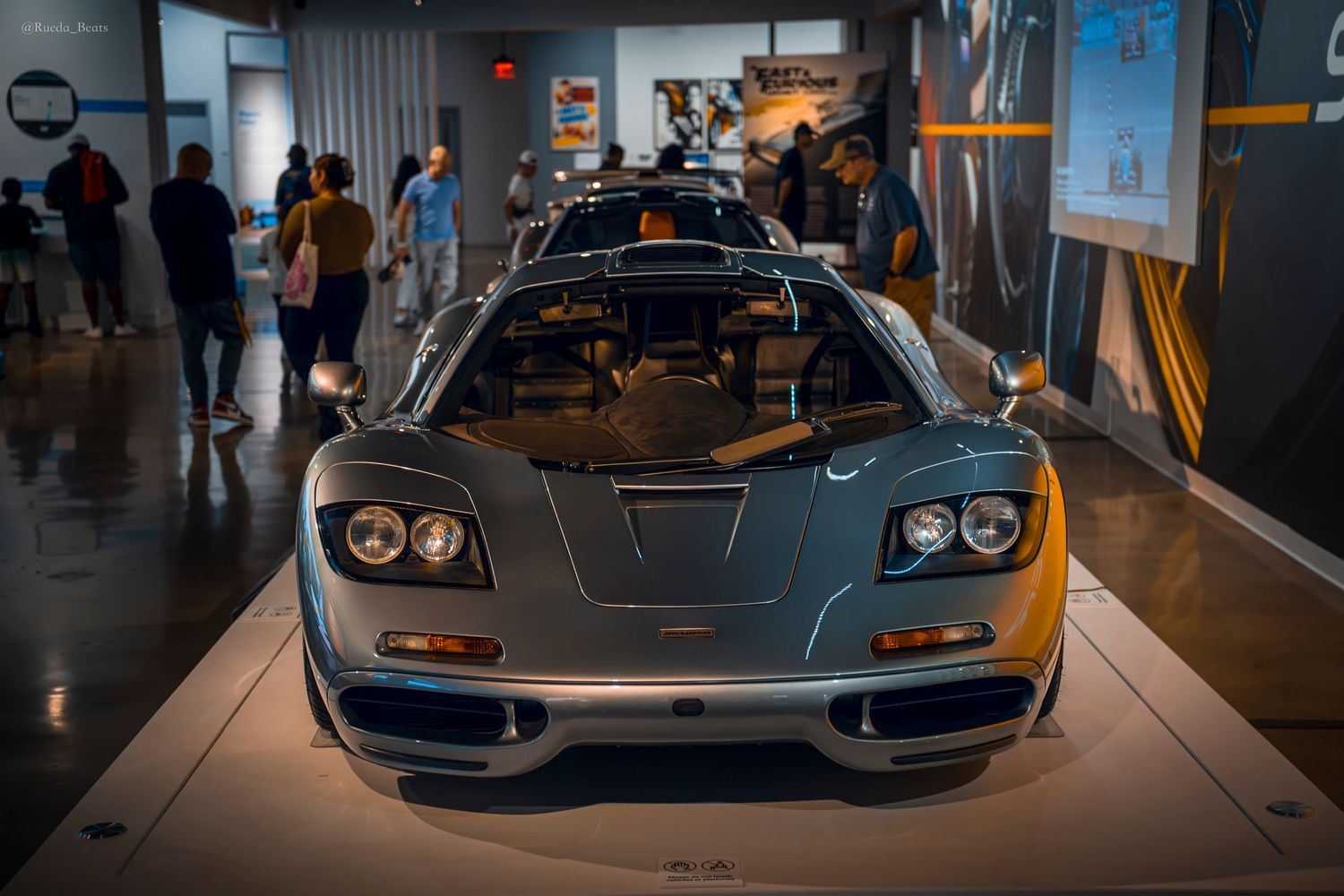 Silver McLaren F1 supercar on display at an automotive museum with visitors in background.