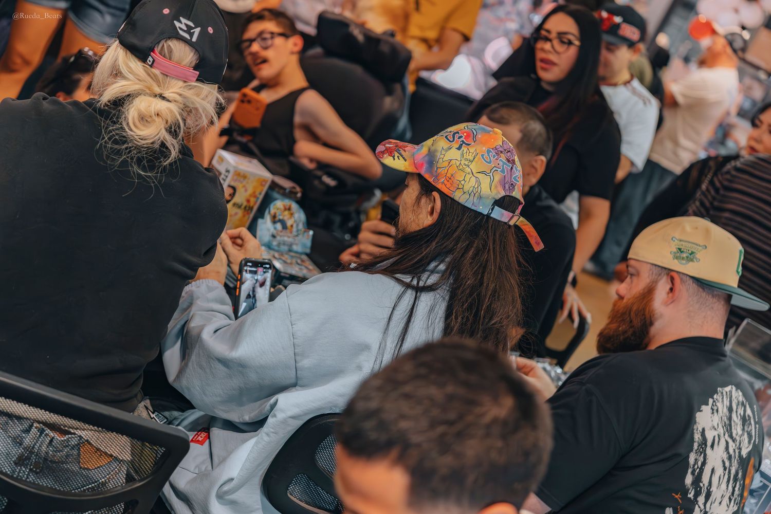 People seated at trading card event with colorful cap collector in focus.