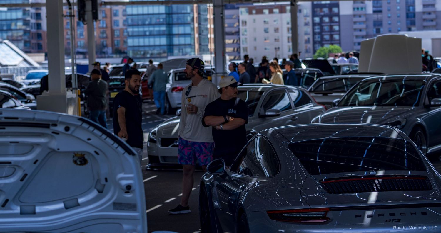 Car enthusiasts browse a Porsche-filled parking lot at an urban automotive meetup event.