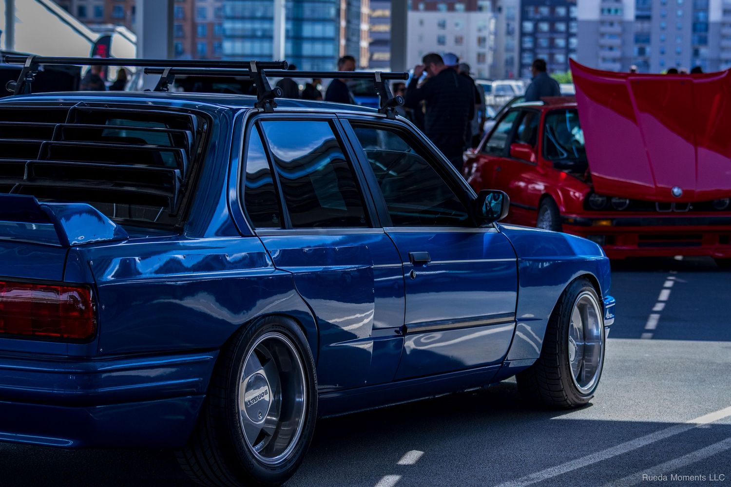 A customized blue sedan with lowered suspension and chrome wheels at an outdoor car show.