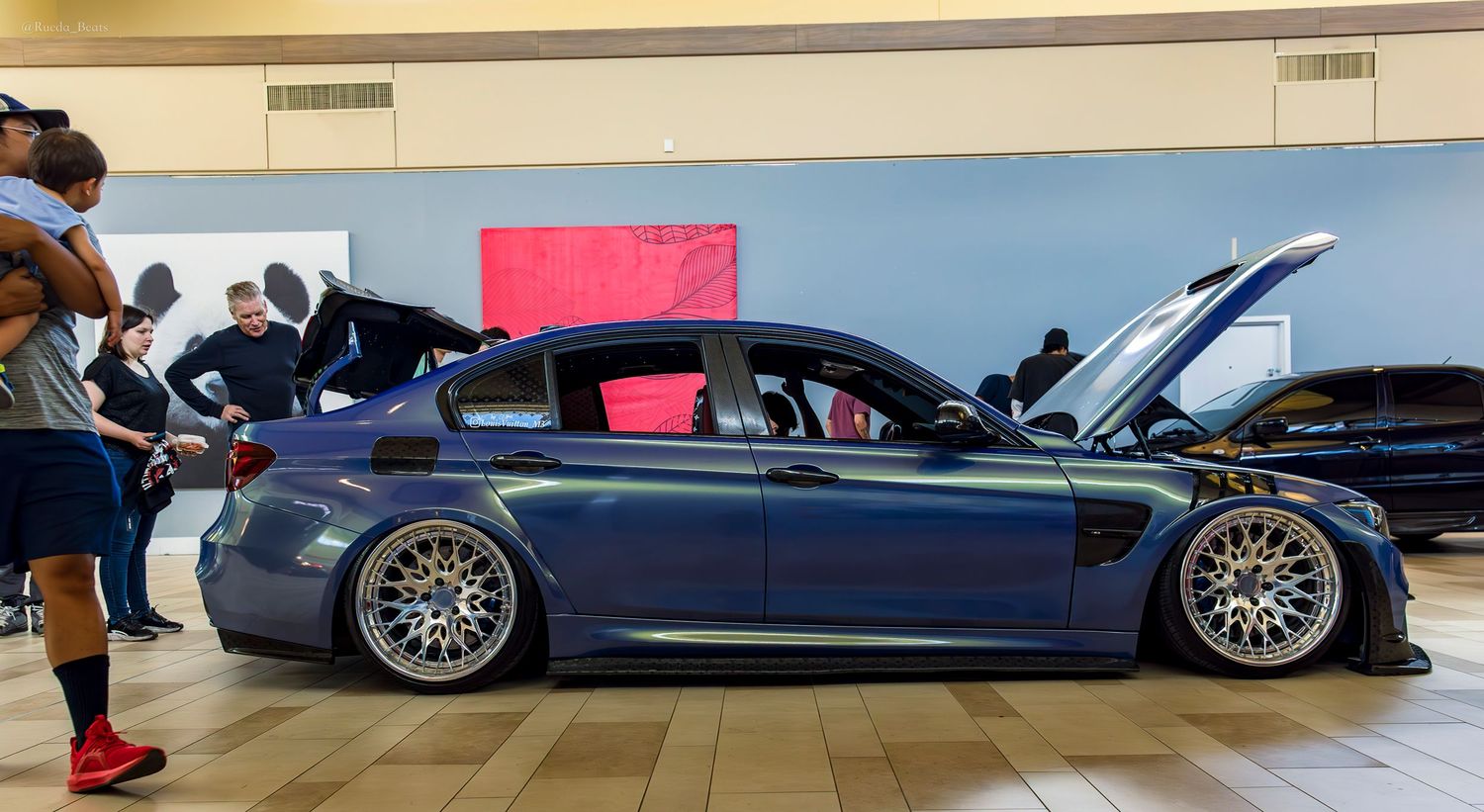 A lowered blue BMW M3 on large chrome wheels displayed at an indoor car show.