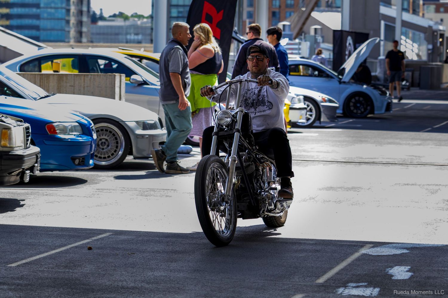 Rider on a classic chopper motorcycle cruising through a busy outdoor car show venue.