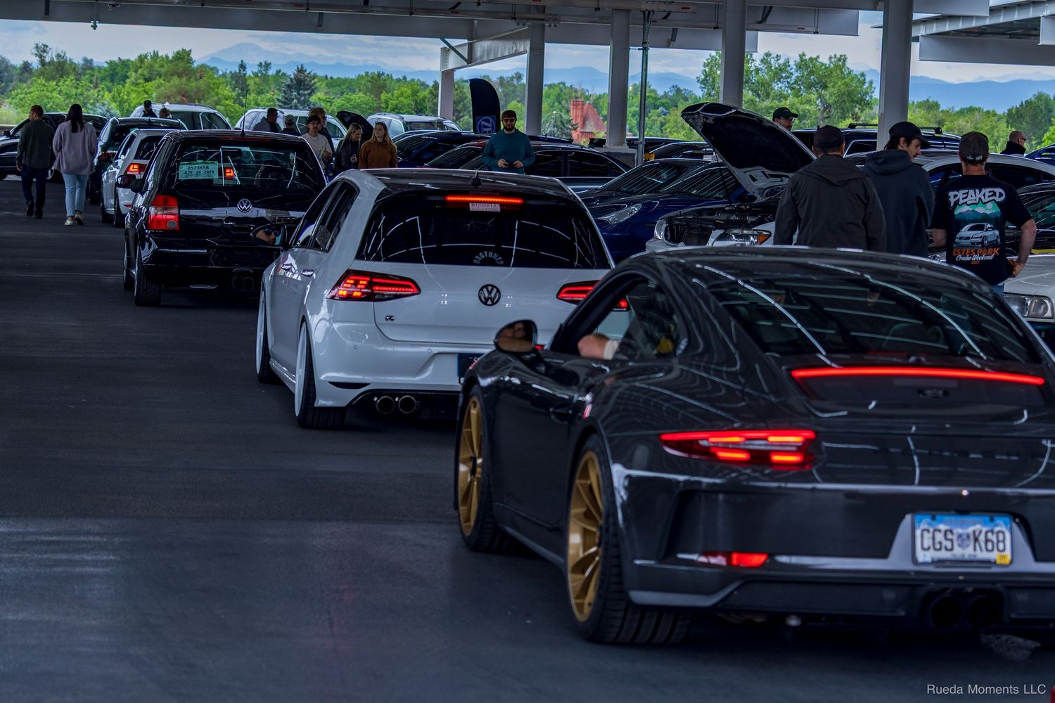 A lineup of modified cars including a VW Golf R and Porsche at an automotive meet in a parking garage.