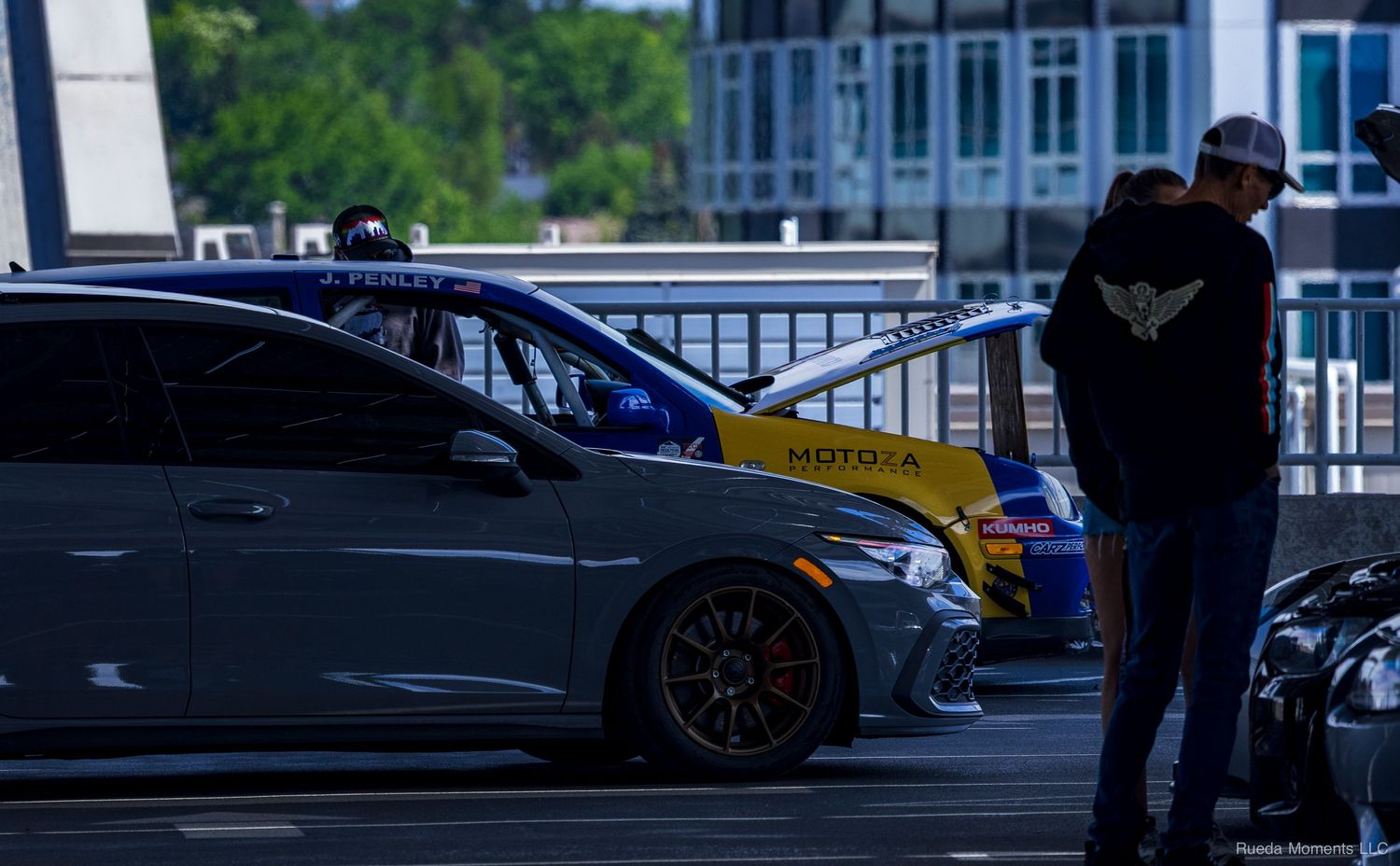 Two people in hoodies examining a blue performance car with yellow accents at an outdoor car show.