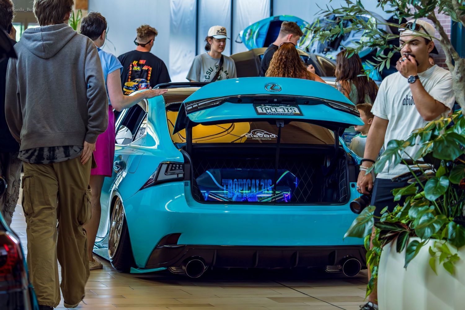 Teal lowered Lexus IS with open trunk audio display surrounded by spectators at car show.