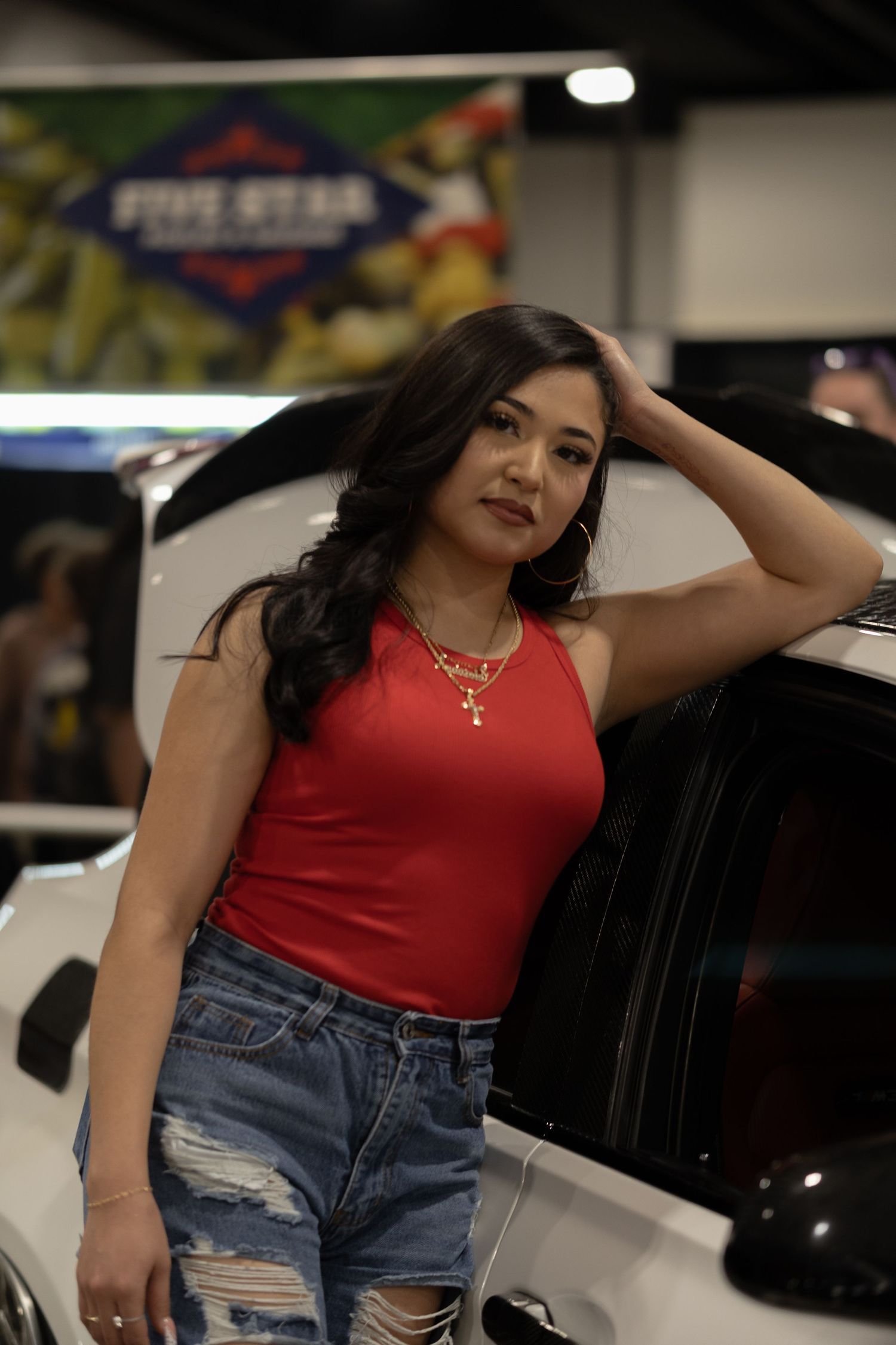 Woman in red halter top and ripped jeans posing beside a white car at an auto show.
