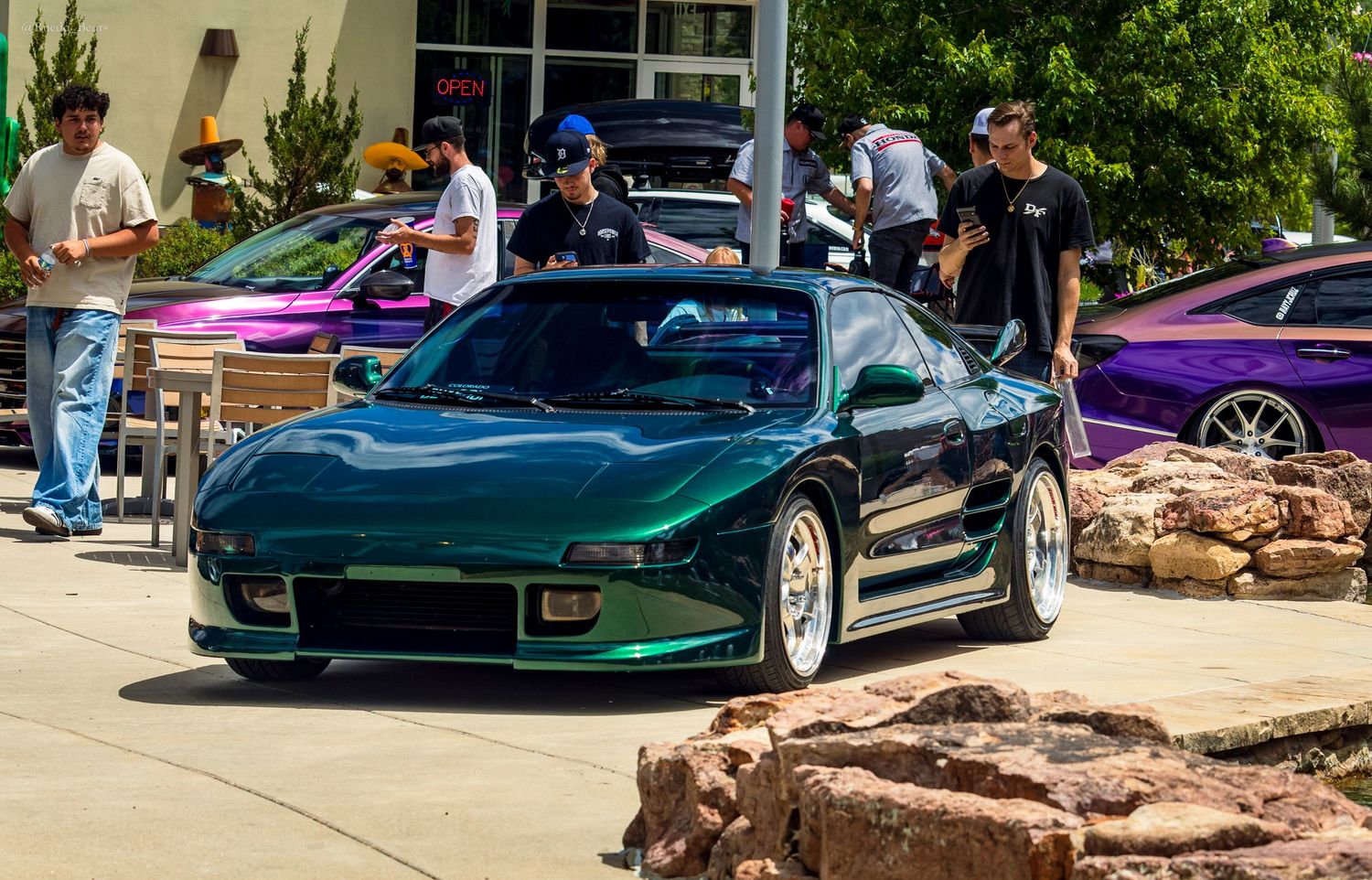 Green Mazda RX-7 with custom wide body kit at an outdoor car enthusiast meet.
