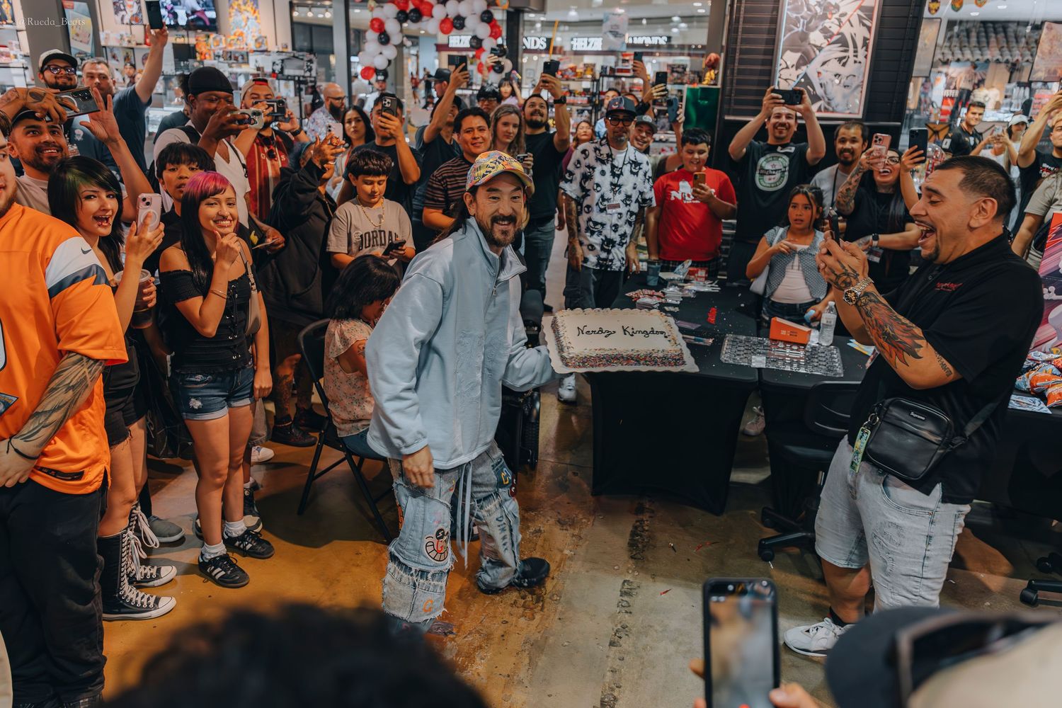 Man holding birthday cake surrounded by cheering crowd at indoor car event.