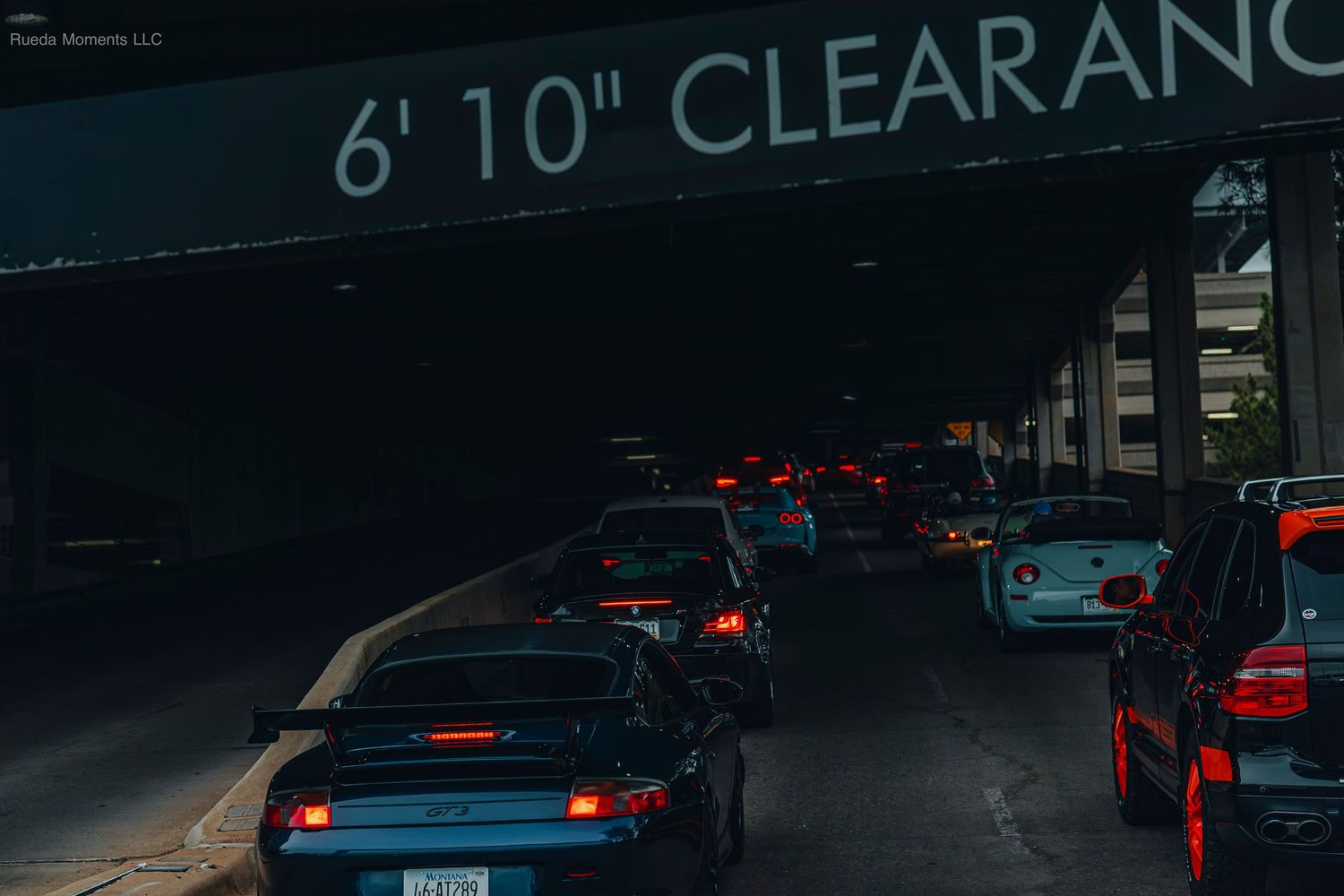 Line of cars including a Porsche 944 passing under a 6 foot 10 inch clearance sign in a garage.