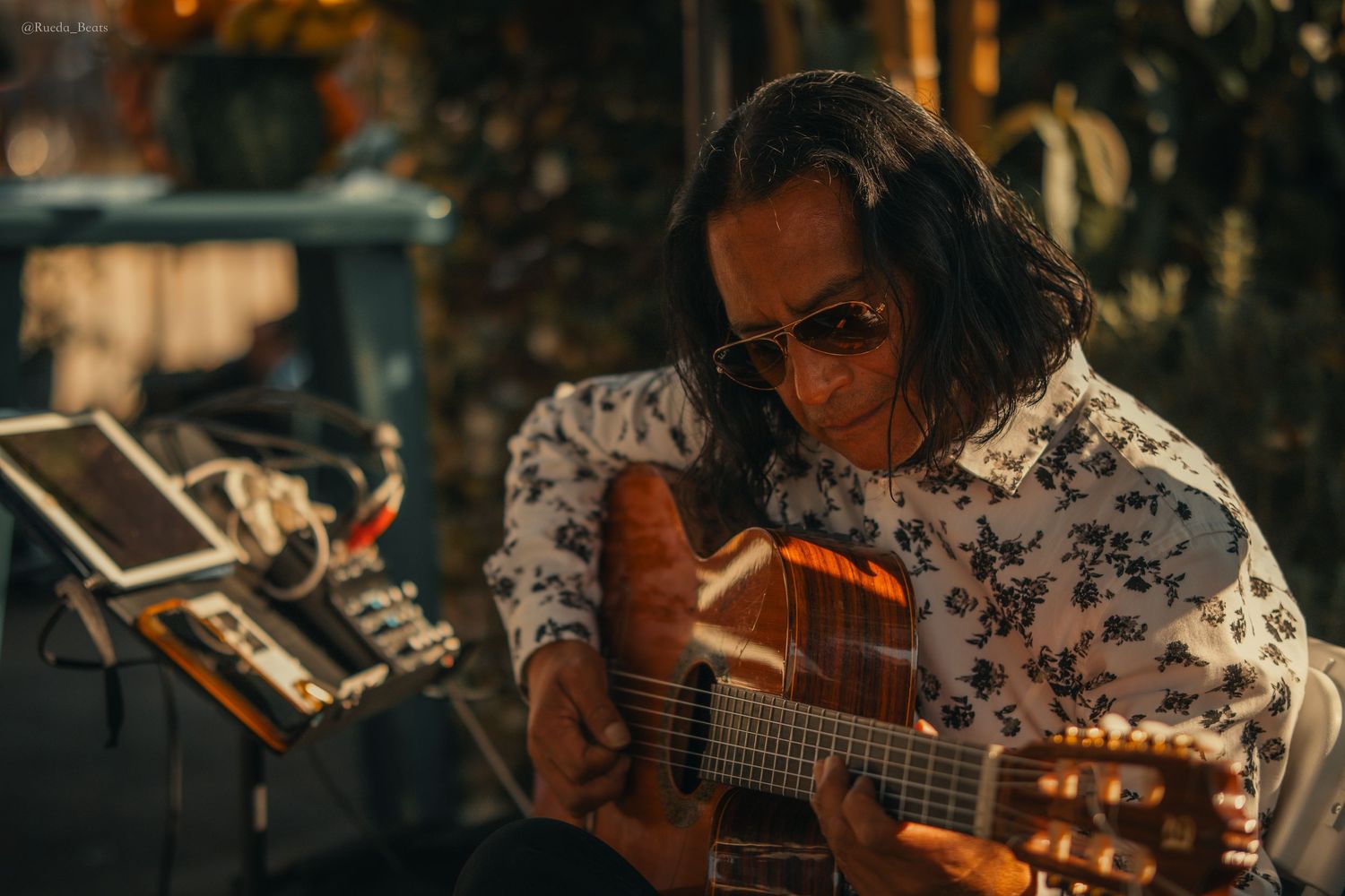 Musician with long hair and sunglasses playing acoustic guitar outdoors in warm golden light.