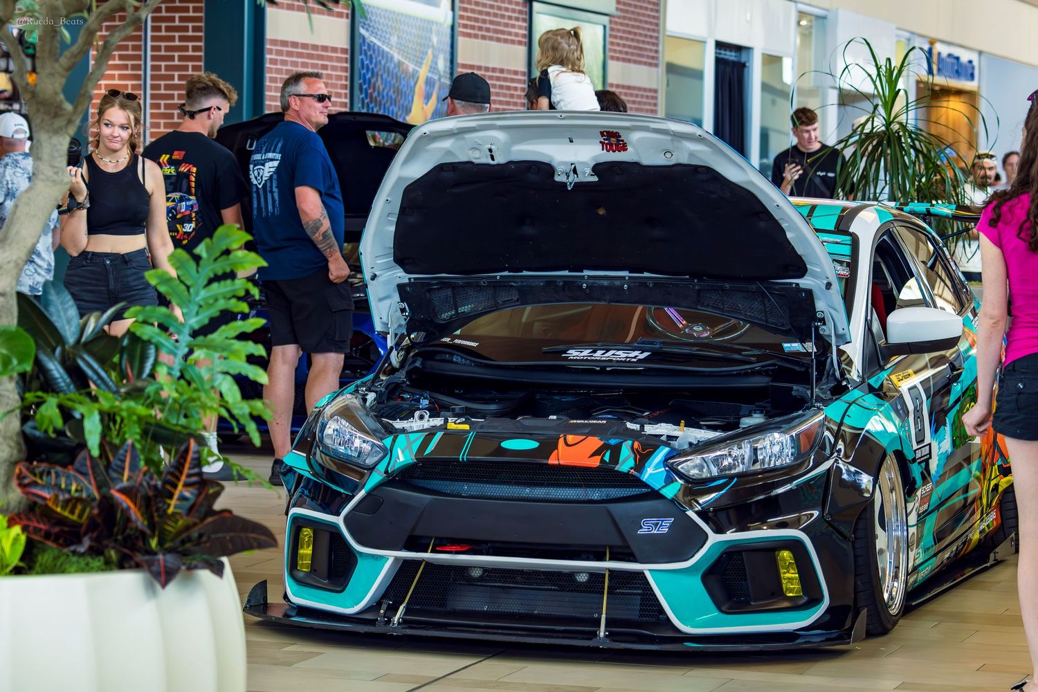 Black and teal widebody Ford Focus RS with open hood at busy outdoor car show event.